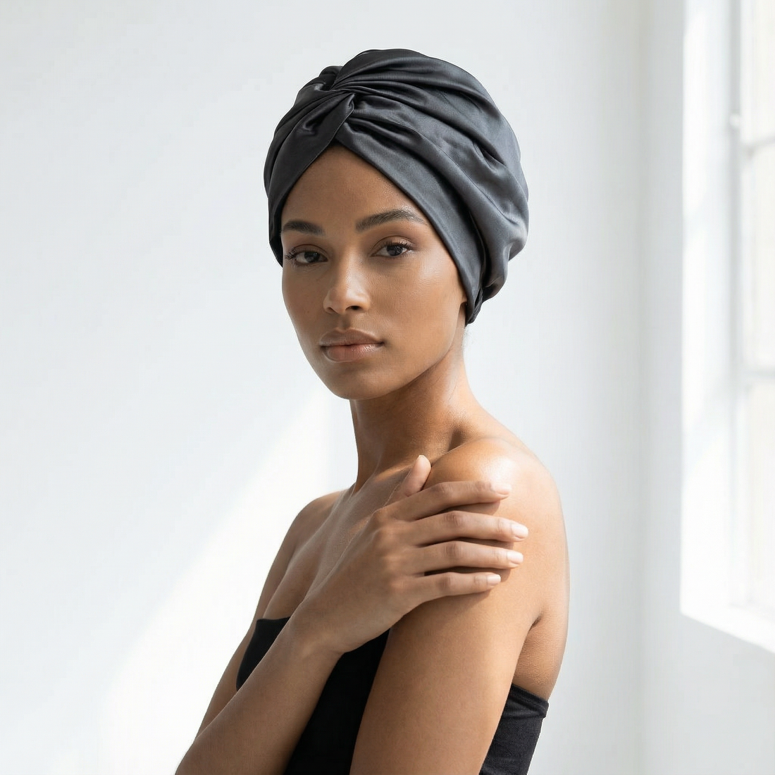 Woman wearing a black silk hair bonnet standing by a window.