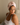 Woman wearing a light pink silk hair bonnet sitting on a bed looking to the side. 