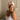 Woman wearing a light pink silk hair bonnet sitting on a bed looking to the side. 