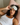 Woman with curly hair lying on a bed wearing a light gray sculpted silk sleep mask on her forehead while laying on a light pink silk pillowcase.