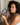Woman with curly hair holding a dark gray silk sleep mask showing the sculpted eye contours that protect lashes while sitting on a bed. 