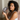 Woman with curly hair holding a dark gray silk sleep mask showing the sculpted eye contours that protect lashes while sitting on a bed. 