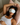 Woman with curly hair lying on a bed wearing a light gray sculpted silk sleep mask and resting on a light pink silk pillowcase, eyes covered, relaxed in soft natural light.