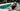 A woman in a black one-piece swimsuit reclines on a white poolside lounge chair with her eyes covered by a champagne colored ZILK Sleep mask, next to a swimming pool with an iced coffee on a side table.