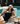 A woman reclines on a white poolside lounge chair wearing a black one-piece swimsuit and a champagne colored ZILK Sleep mask. A glass of iced coffee sits on a wooden side table next to her, with a swimming pool and outdoor patio in the background.